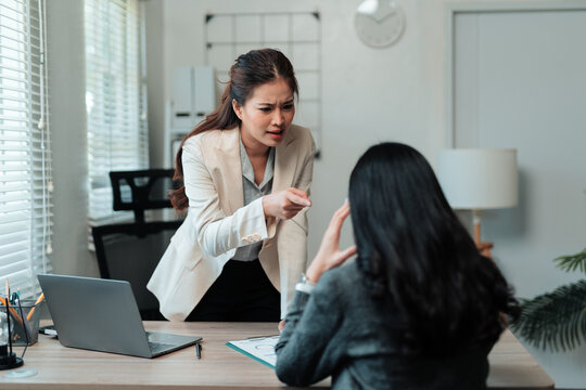Asian female boss yelling and pointing finger at upset employee during workplace conflict, showing stress and confrontation