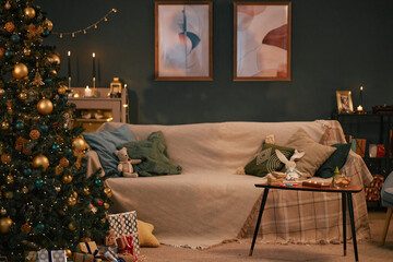 Living room decorated for Christmas with Christmas tree, wrapped gifts, plush toy on sofa, candles and framed abstract art on wall, cozy holiday setting with copy space