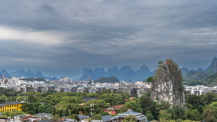 Fototapeta premium Beautiful urban landscape. Multi-storey buildings, green vegetation. At the top of a high sheer cliff, a pavilion with a curved roof is visible. Mountains against the sky and clouds. China. Guilin