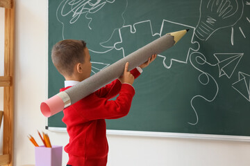 Cute little schoolboy with large pencil drawing on chalkboard in classroom
