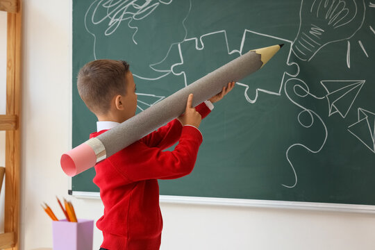 Cute little schoolboy with large pencil drawing on chalkboard in classroom