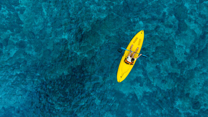 Aerial view woman bright yellow kayak with in the vast expanse of crystal clear turquoise water. The seabeds rocky texture is visible beneath the surface, concept marine environment
