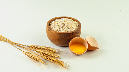 Dry oatmeal in a wooden bowl and a broken egg beside it on a white background