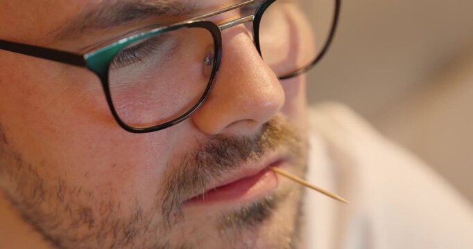 A close-up of a man&rsquo;s face with a toothpick in his mouth, showing a relaxed and thoughtful expression under warm indoor lighting.