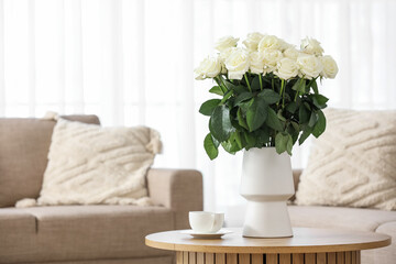 Vase with white roses and cup of coffee on table in living room