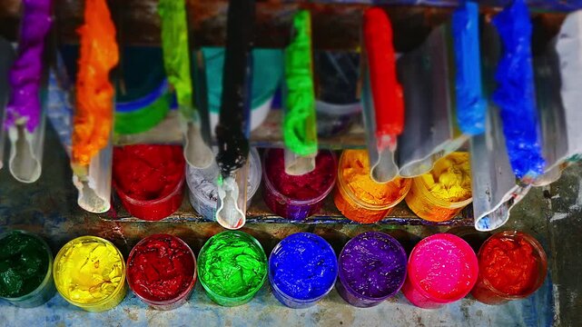 On a wooden workbench in a screen printing shop, several plastisol ink cans are open, revealing rich tones soft pastels, strong primaries, and deep shades. A wooden spoon and trowel rest nearby.