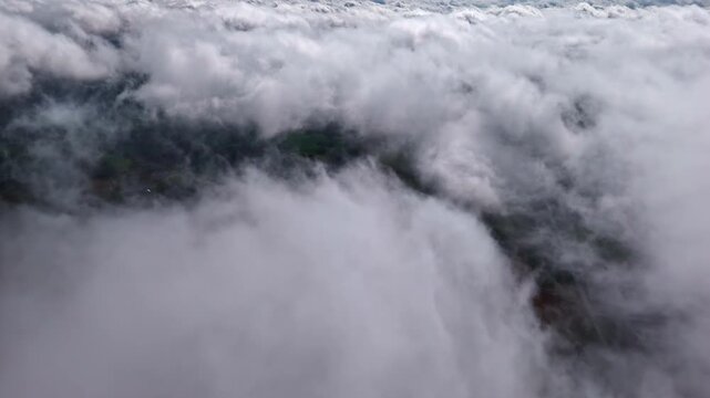 High altitude aerial view flying forward over white clouds with patches of green fields and land in Latvia visible below