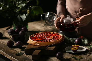 Woman with tasty plum pie, spatula, teapot of tea and cups at wooden table against dark background