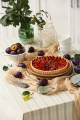Plate with tasty plum pie, cups and fresh fruits on table in kitchen