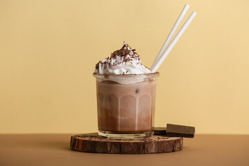 Glass of cold cocoa drink with whipped cream and chocolate on brown table against yellow background
