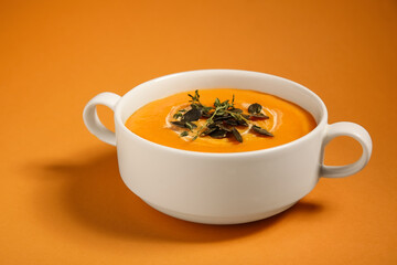Pot of delicious pumpkin cream soup with seeds and herbs on orange background, closeup