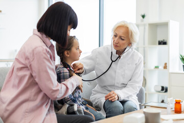 Obraz premium A female doctor is using a stethoscope to listen to a young girl's heart while the mother sits beside them. The scene takes place in a doctor's office.