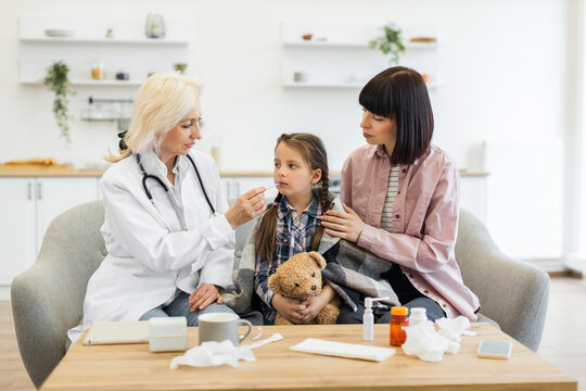 A doctor examines a young girl with a thermometer while the mother comforts her, showing care and concern in a home setting. - Powered by Adobe