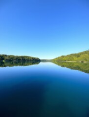 Obraz premium Calm Lake Scene: The image presents a pristine lake under a vast, cloudless blue sky, with lush greenery gracing its banks. The tranquil waters mirror the landscape.