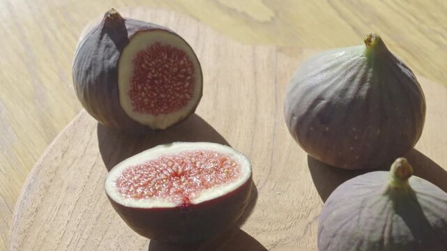 Fresh figs sit on a wooden board, with one fig sliced open to showcase its vibrant red pulp and tiny seeds. Natural sunlight highlights the fruit's texture and colors.