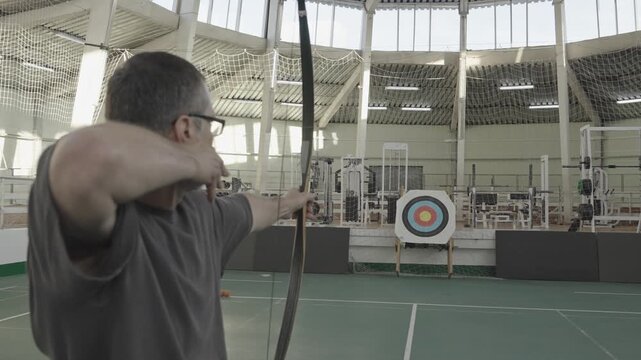 An individual practices archery indoors at a sports facility, focusing on form and precision while aiming at the target. The environment features a well-equipped training area.