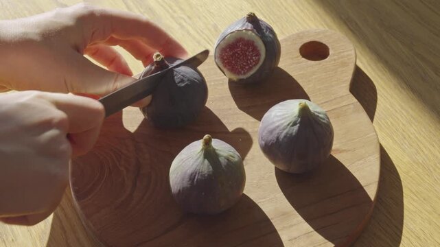 A hand is slicing ripe figs on a wooden cutting board in bright natural light. The focus is on the juicy insides and perfect texture of fresh fruit. Enjoy this simple preparation method.