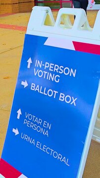 Pedestrians move past while a ballot drop-off sign with bold arrows shows Spanish text Votar en Persona and Urna Electoral, highlighting civic responsibility, safety, democracy, freedom. Vertical