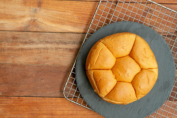 A top-down flat lay of a round, segmented bread roll on a dark slate plate. The plate sits on a wire cooling rack, all set against a warm, brown wooden table providing copy space.