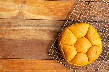 A top-down flat lay of a round, segmented bread roll on a metal cooling rack. The rack is placed on a brown wooden table, which provides ample copy space on the left.