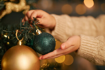 Child hands decorating Christmas tree with glittery ornaments, gently placing blue bauble on...