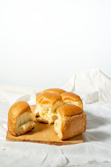 Torn pieces of a soft, golden-brown bread roll rest on a wooden board, showing the fluffy interior. The setting is on a white cloth with a plain white background providing copy space.