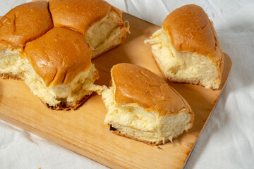 A close-up of a soft, golden-brown bread roll torn into several pieces, revealing the fluffy interior, all placed on a wooden cutting board over a white cloth.
