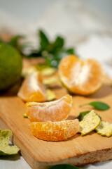 Peeled orange segments are stacked on a wooden cutting board. Green peels, leaves, and a halved orange are scattered nearby, with a shallow depth of field.