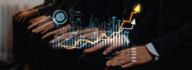 Business professionals are seated at a conference, with digital graphs and charts illustrating...