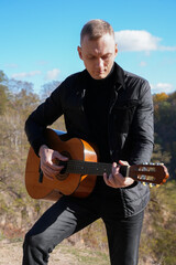 Beautiful blonde man playing acoustic guitar and singing relaxing enjoying outdoors on sunny autumn day. Artist learning and practicing playing musical instrument. Fall leaves autumnal weather 