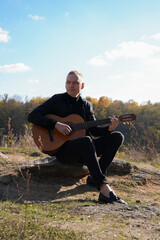 Blonde man playing string guitar outdoors in autumn forest. Concept of sound therapy, mental health and wellness rituals. Calmness tranquility audio-sensory practices. Aura farming energy holistic