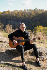 Beautiful blonde man playing acoustic guitar and singing relaxing enjoying outdoors on sunny autumn day. Artist learning and practicing playing musical instrument. Fall leaves autumnal weather 