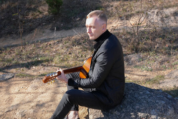 Blonde man playing string guitar outdoors in autumn forest. Concept of sound therapy, mental health and wellness rituals. Calmness tranquility audio-sensory practices. Aura farming energy holistic