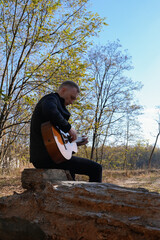 Blonde man playing string guitar outdoors in autumn forest. Concept of sound therapy, mental health and wellness rituals. Calmness tranquility audio-sensory practices. Aura farming energy holistic