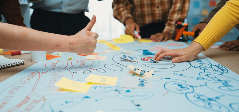 A dynamic workshop scene showcasing multiple hands engaged in writing and planning on a large paper with sticky notes, highlighting teamwork and creative thinking. SACTR