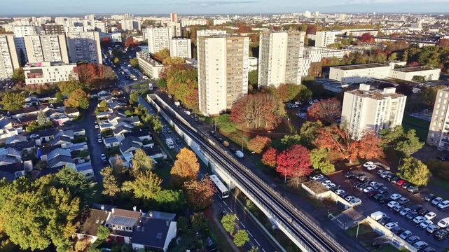 Drone flying above La Poterie metro station in Rennes on a sunny day, showing the elevated tracks, nearby roads with buses and cars, parking lots and residential buildings.
