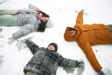 Caucasian man, woman and child lying on snow making snow angels, smiling and looking up, winter outdoor activity, family enjoying time together