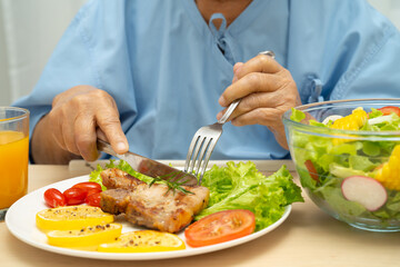 Asian senior woman patient eating pork chop stake and vegetable salad for healthy food in hospital.