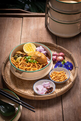 Beautiful still life of traditional Northern Thai Khao Soi chicken curry noodles served in an enamel bowl on a wooden table, with crispy noodles, shallots, lime, chili oil.