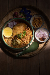 Beautiful still life of traditional Northern Thai Khao Soi chicken curry noodles served in an enamel bowl on a wooden table, with crispy noodles, shallots, lime, chili oil.