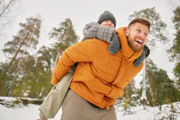 Caucasian man giving piggyback ride to Caucasian child outdoors in snowy forest, both smiling and enjoying winter activity, trees and snow visible in background