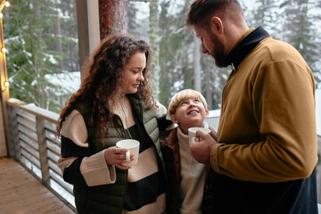 Caucasian woman, man, and child standing together on outdoor wooden terrace holding mugs, child looking up at man, winter forest visible in background