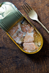 Open tin can of salmon fish fillet in its own juice and metal fork on wooden table background, closeup, top view