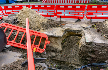 Street excavation with red safety barriers, groundworks for underground infrastructure repair, urban network modernization during road maintenance in public city area