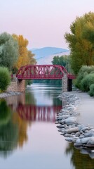 Obraz premium Curved Red Metal Truss Bridge Spanning Calm River Reflecting Soft Sky and Lush Green Trees During Golden Hour