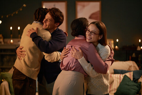 Two young adult women and two young adult men, including Black and Caucasian individuals, hugging each other in warmly lit living room, expressing happiness and connection