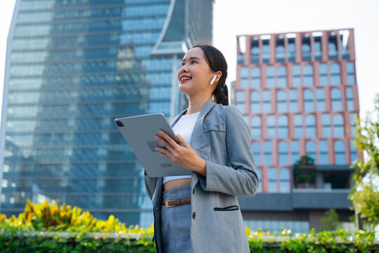 successful young Asian businesswoman in a stylish gray suit standing outside a modern glass office building, holding a digital tablet. Concept of corporate success, urban professional, and digital tec