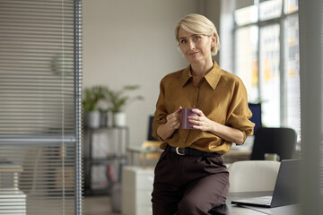 Portrait of middle aged Caucasian woman standing in modern office holding coffee mug, smiling...