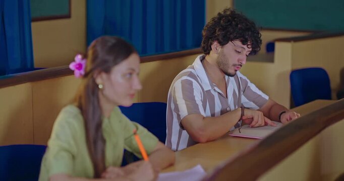 Indian young adult gen z male man lady female hold pencil writing notes do study sitting desk table class room place. Happy pupil boy learn course share doubt raise hand new day inside college hall