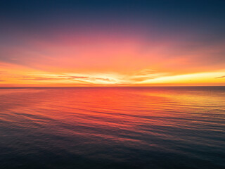 Vibrant Stunning Sea Sunset Ocean Sunrise Over Calm Ocean water At Dusk With Reflective Ripple surface of Red And Orange Waves, Aerial view